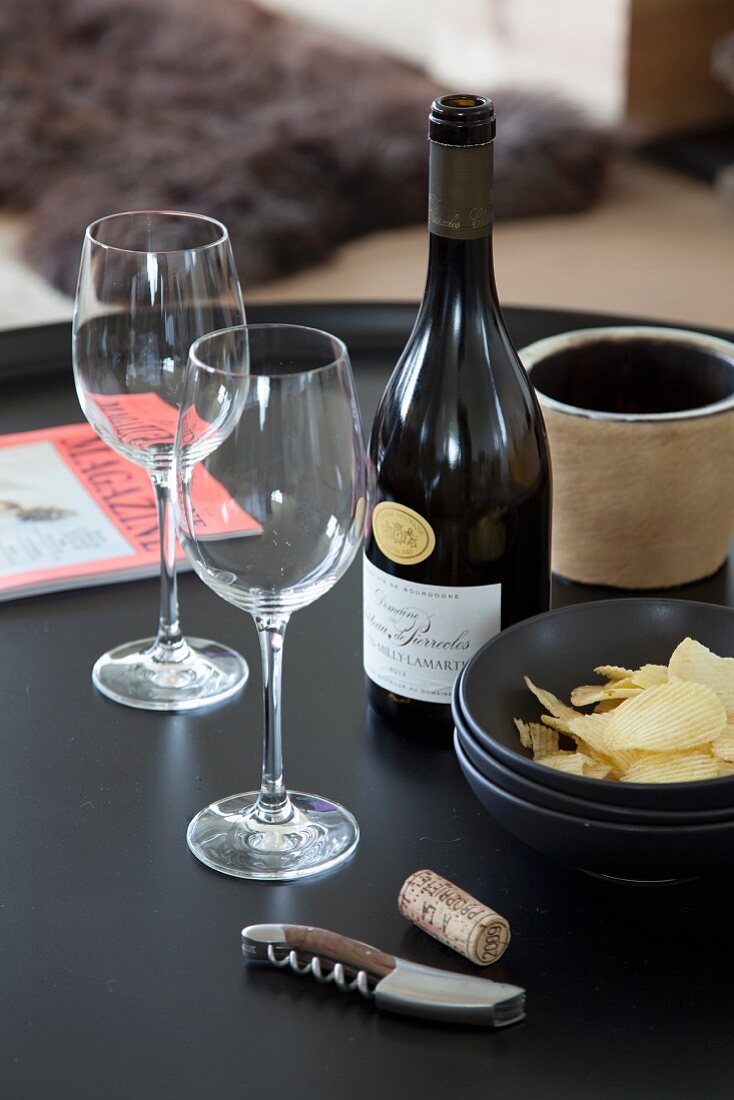 Bottle of fine wine, two wine glasses, bowl of crisps and corkscrew on black tray table