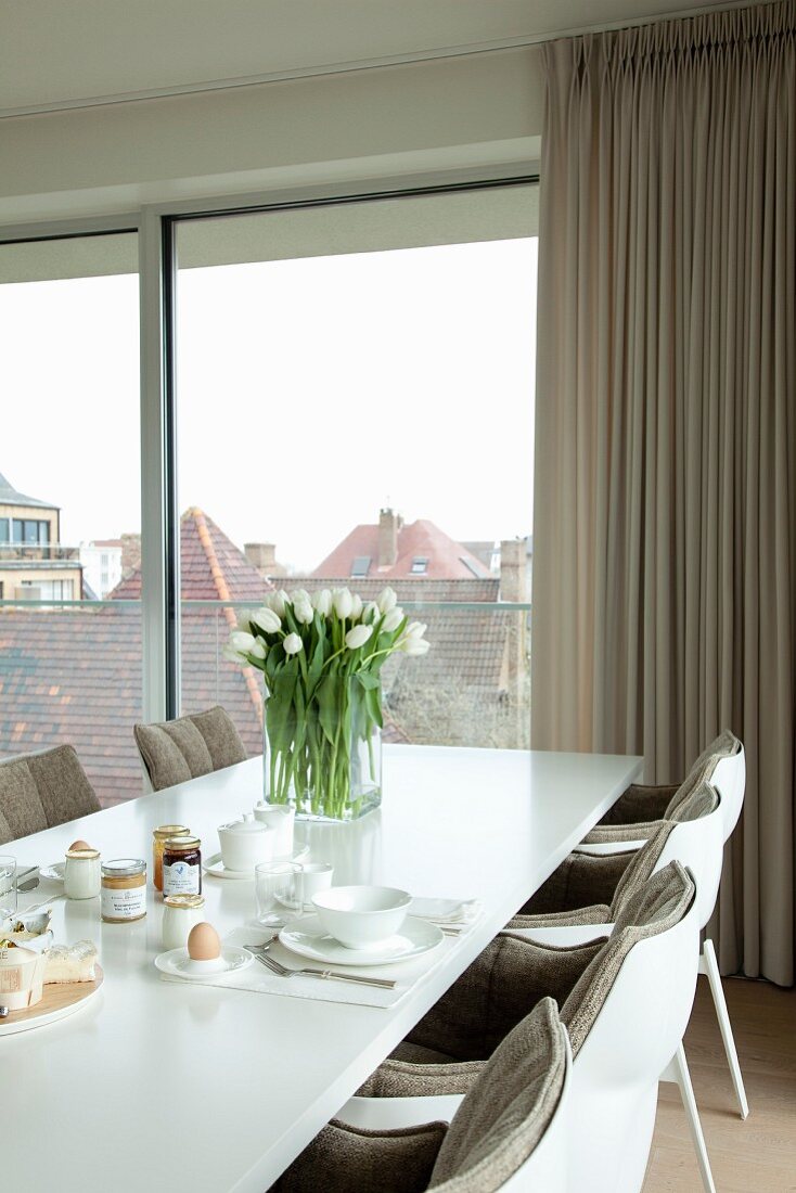 White dining table set for breakfast with vase of white tulips and upholstered designer chairs next to glass wall