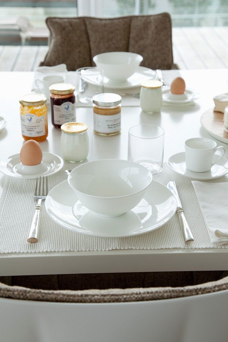Two white breakfast place settings with silver cutlery, boiled eggs and jars of jam