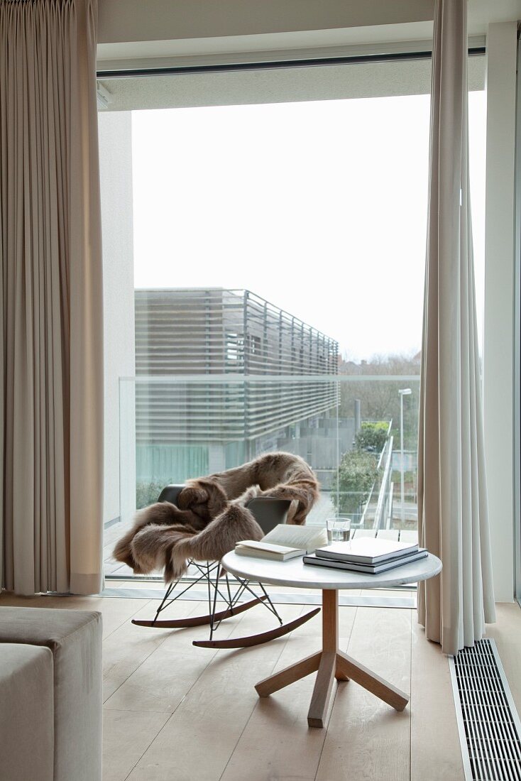 Eames rocking chair, fur blanket, boos on round table, glass wall and floor-length beige curtains in reading corner