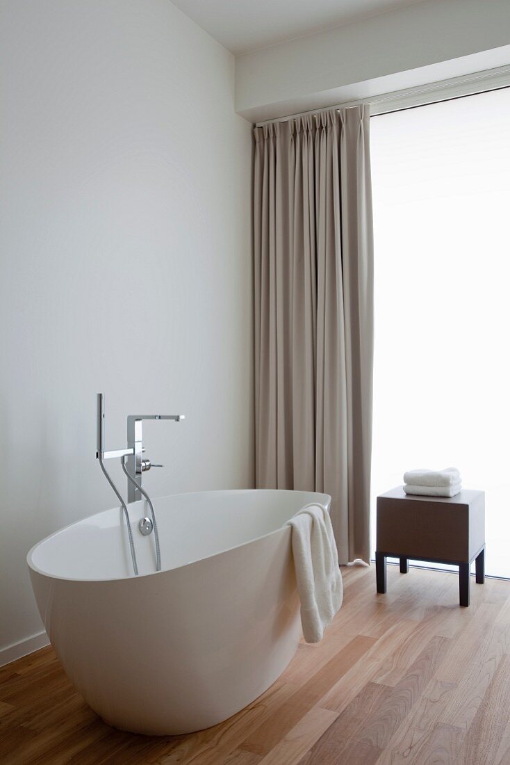 Free-standing white bathtub on teak parquet floor, floor-length beige curtain, stool and frosted glass wall