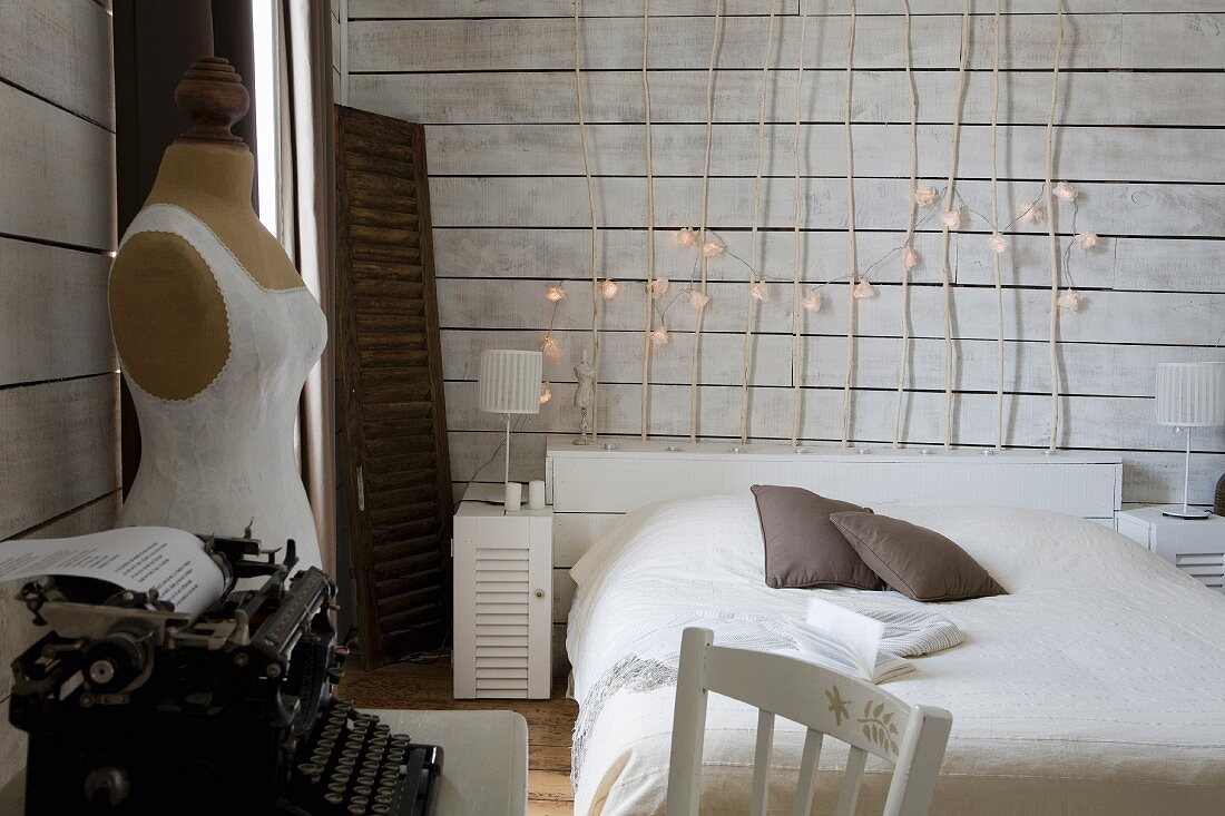 Schlichtes Bett vor Holzablage, darauf Stäbe mit Lampengirlande, seitlich Vintage Schreibmaschine neben Schneiderpuppe, in weißem holzverkleidetem Schlafzimmer