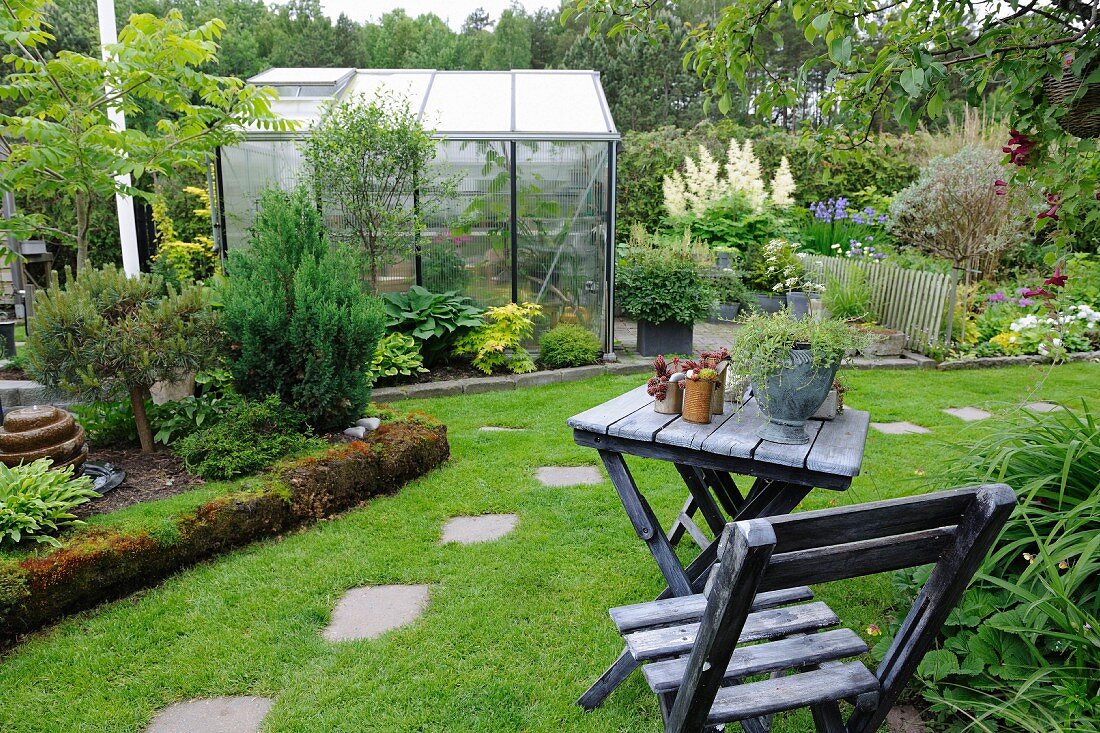 View past weathered wooden table and chair to greenhouse and flowerbeds in summer garden