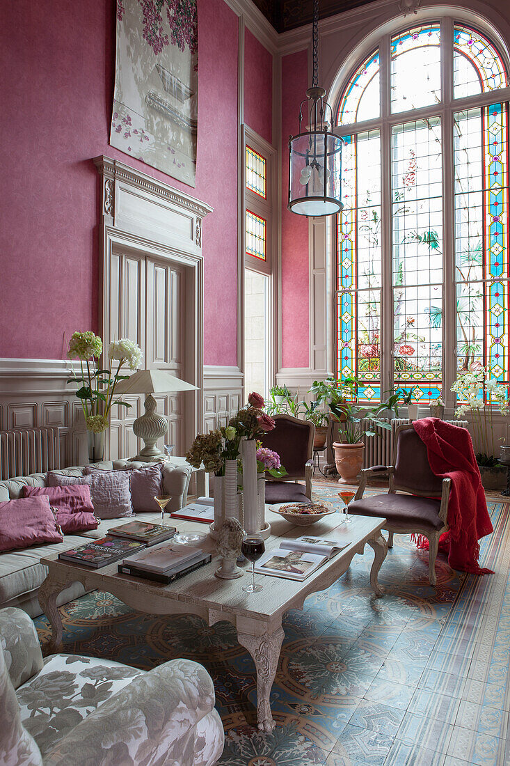 Coffee table and antique seating in grand lounge with high ceiling, dusky-rose wallpaper and floor-to-ceiling window with artistic stained-glass elements