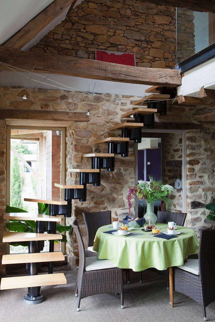 Tisch mit grüner Tischdecke und Rattanstühle vor Wendeltreppe in Wohnraum mit Natursteinwänden