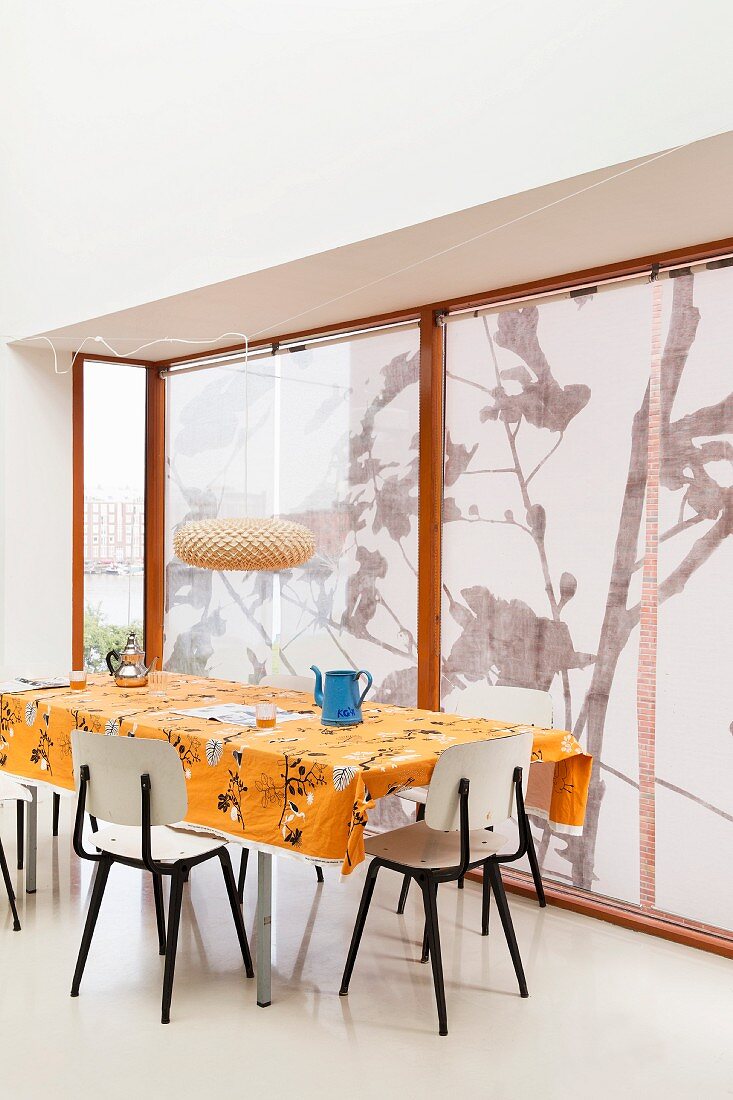 Retro Stühle um Tisch mit orangefarbener Tischdecke vor Fenster mit Rollos
