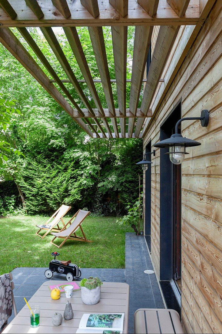 Blick von Terrasse über Tisch auf Liegestühle im Garten, vor Wohnhaus mit Holzlamellen Sonnenschutz