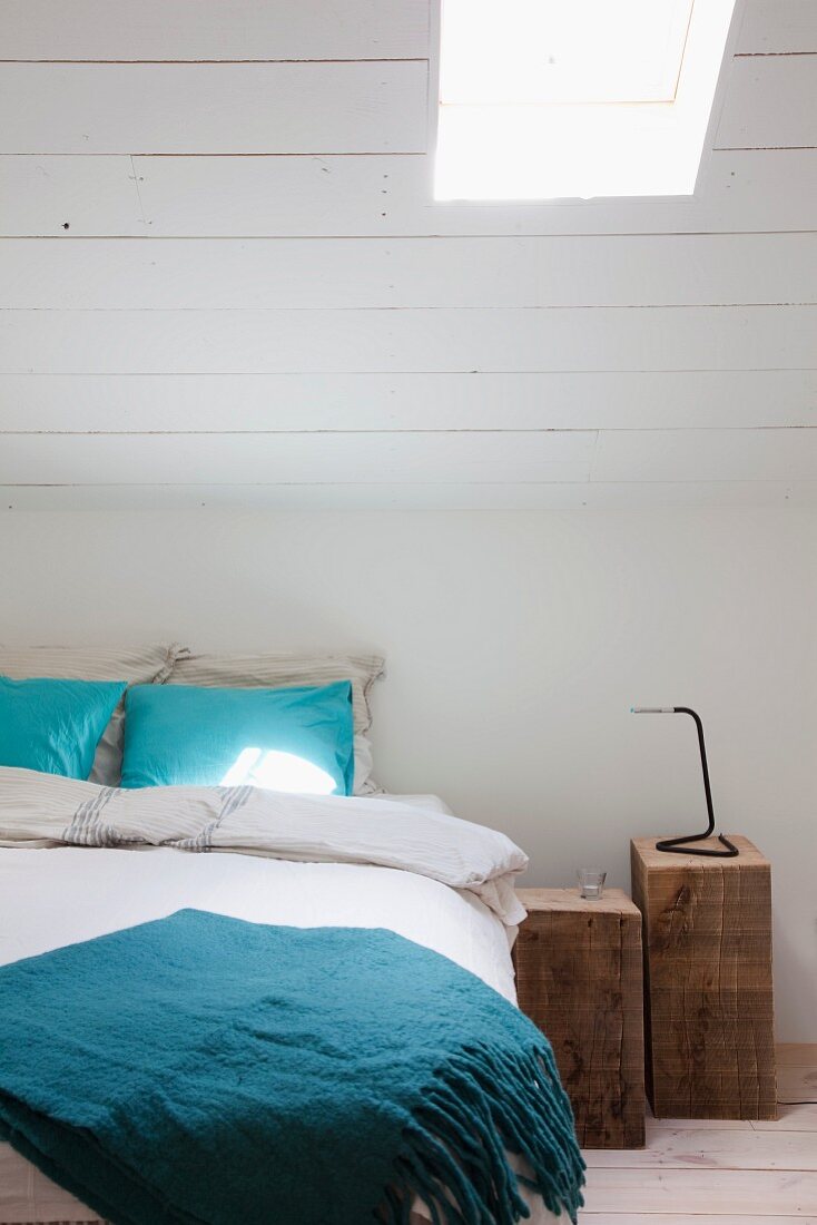 Zwei rustikale Holzblöcke als Nachttische neben Bett und weiß gestrichene Dachschräge mit Dachflächenfenster