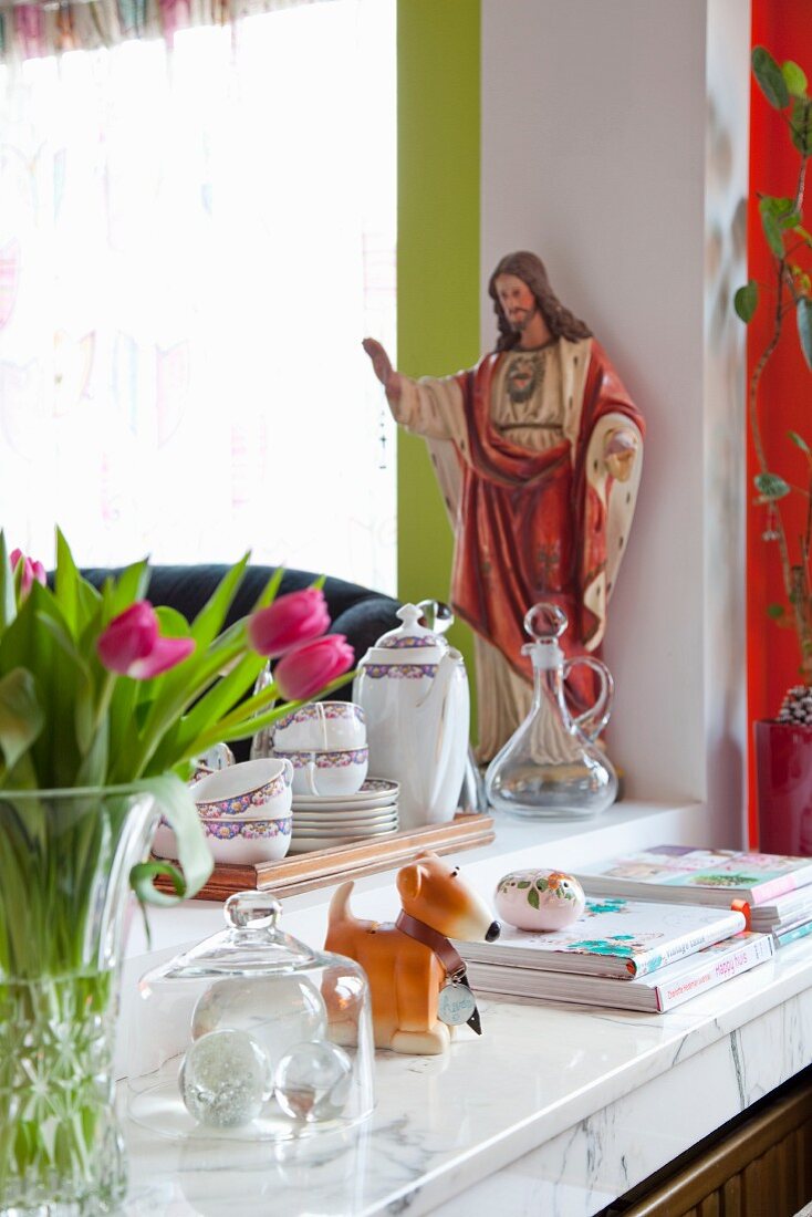 Tulpenstrauss und Bücherstapel auf Ablage vor Durchreiche, darauf Tablett mit Geschirr, im Hintergrund religiöse Figur