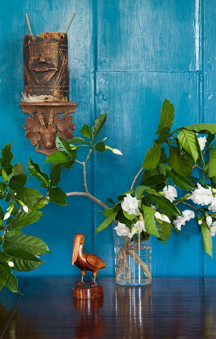 Carved pelican figurine and flowering branches in vase below wooden ethnic carvings on wall painted light blue