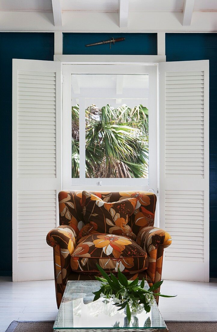 Armchair with brown, retro floral pattern and wicker table with glass top in front of French window with interior folding shutters