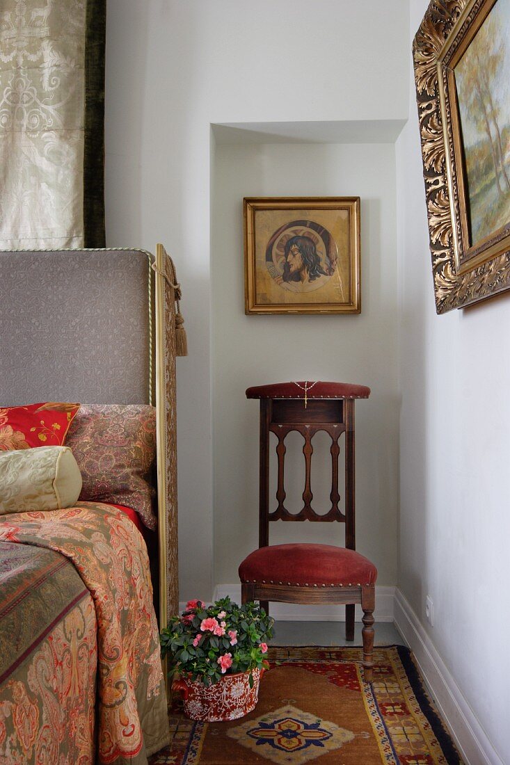 Icon and antique chair in niche next to bed with upholstered headboard and traditionally patterned bed linen