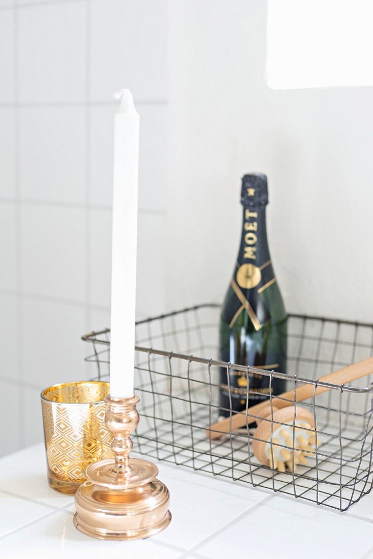 White candles in gilt candlestick and gold tealight holders in front of bottle of Champagne in wire basket