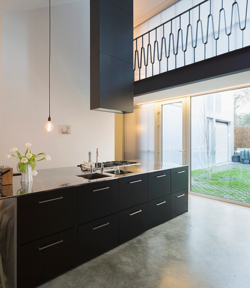 Designer kitchen counter with black drawers and stainless steel handles below extractor hood in minimalist interior; view through floor-to-ceiling window in background