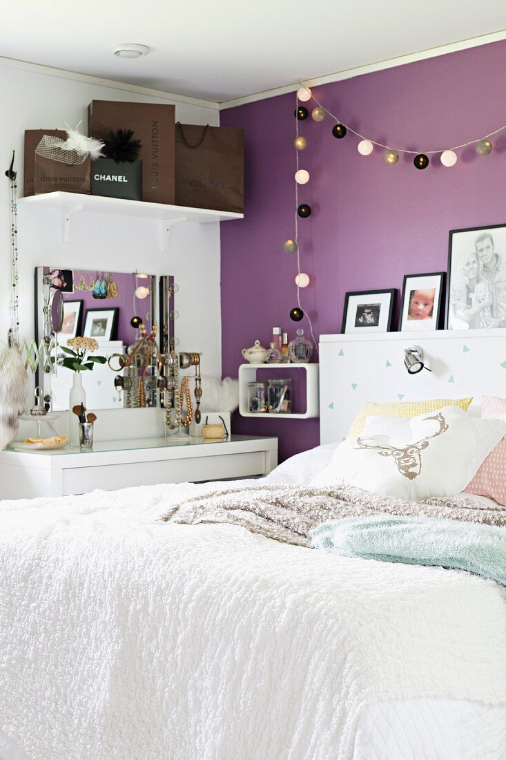Bett mit weisser Tagesdecke in modernem Schlafzimmer, Lampiongirlande an violett getönter Wand aufgehängt