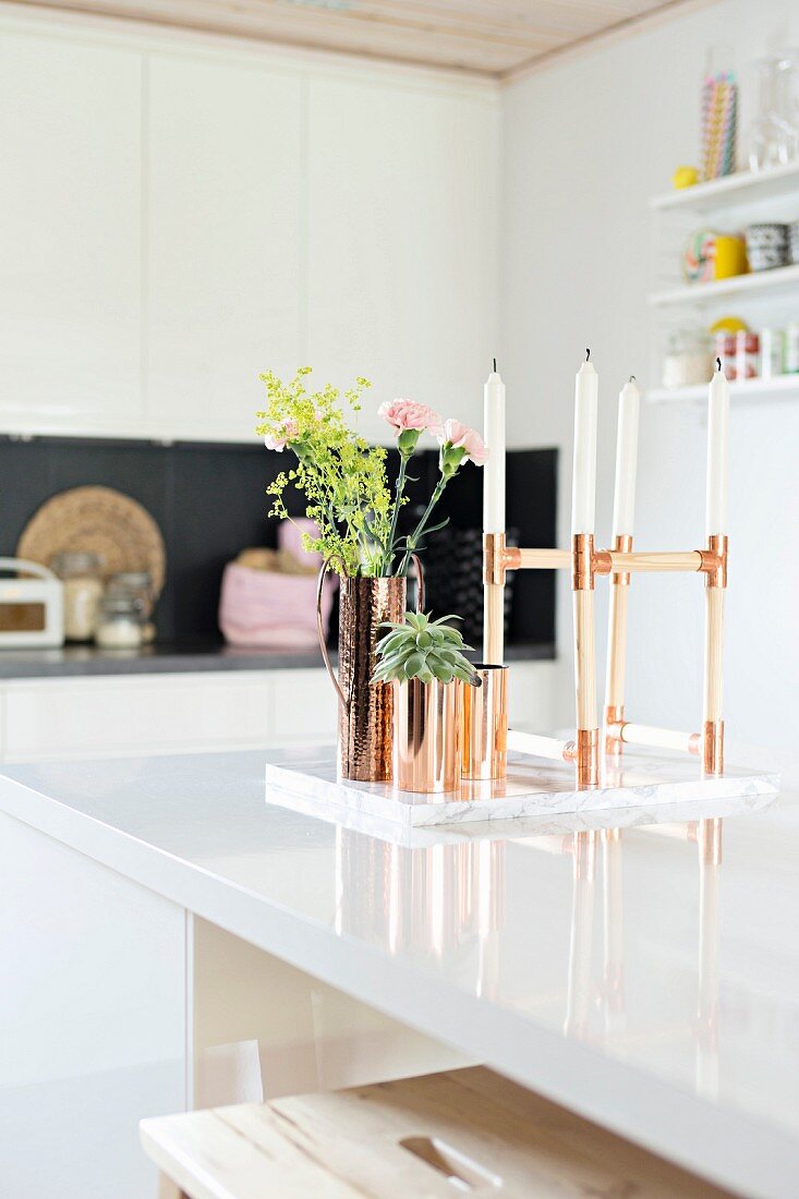 Kerzenständer mit weissen Kerzen und kupferfarbene Behälter mit Blumen und Sukkulente auf weiss lackierter Theke in moderner Küche