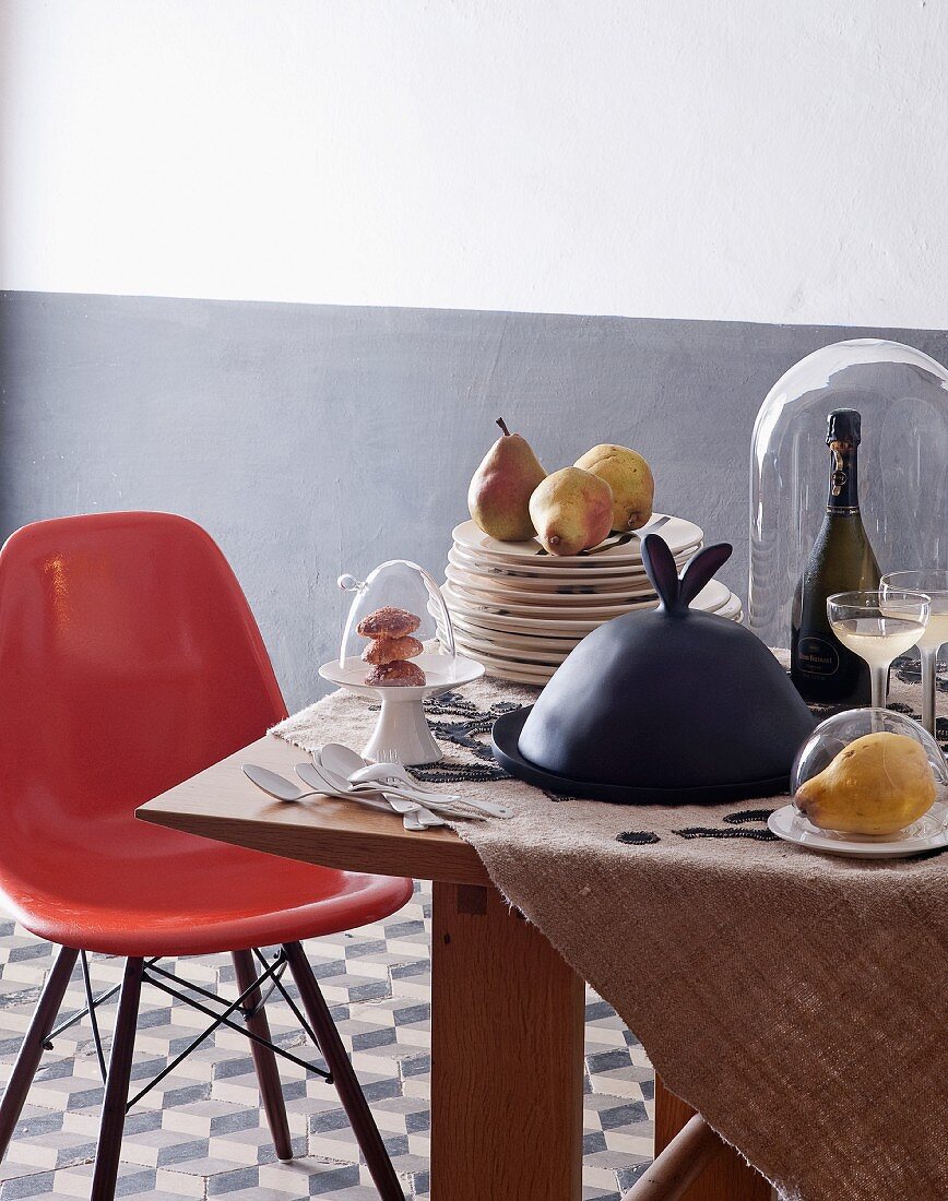 Eames chair at wooden table with stacked plated, food under glass covers and bottle of Champagne