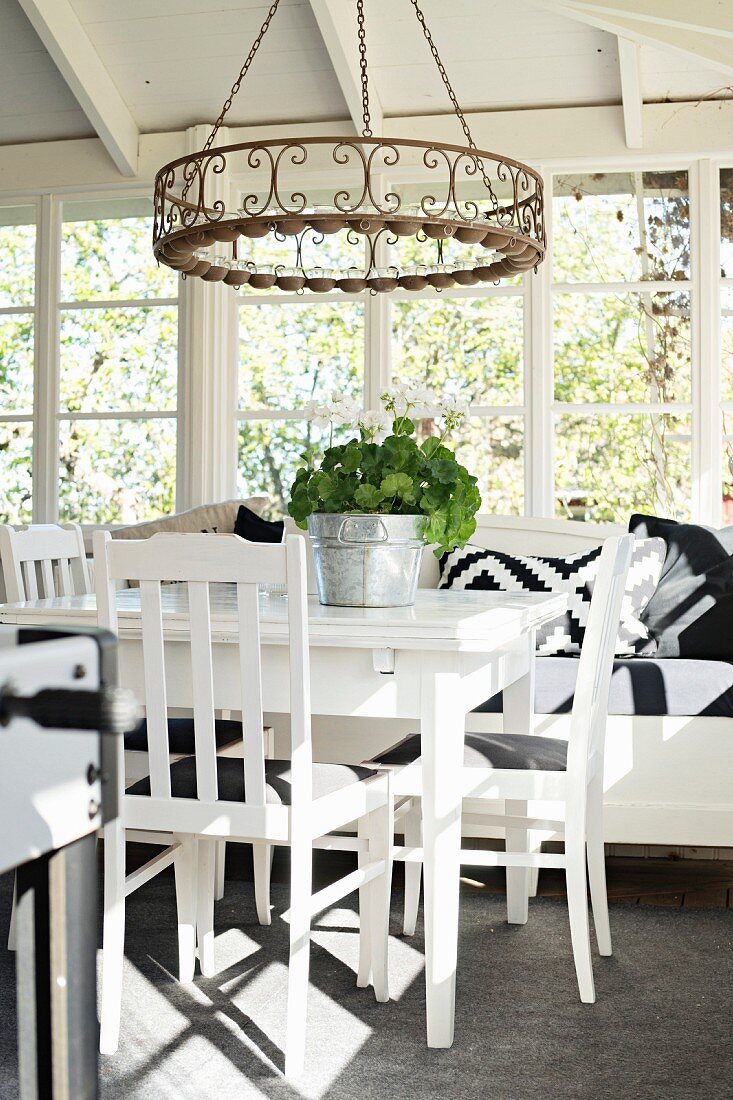 White-painted table, chairs and bench below wrought iron candle chandelier in conservatory