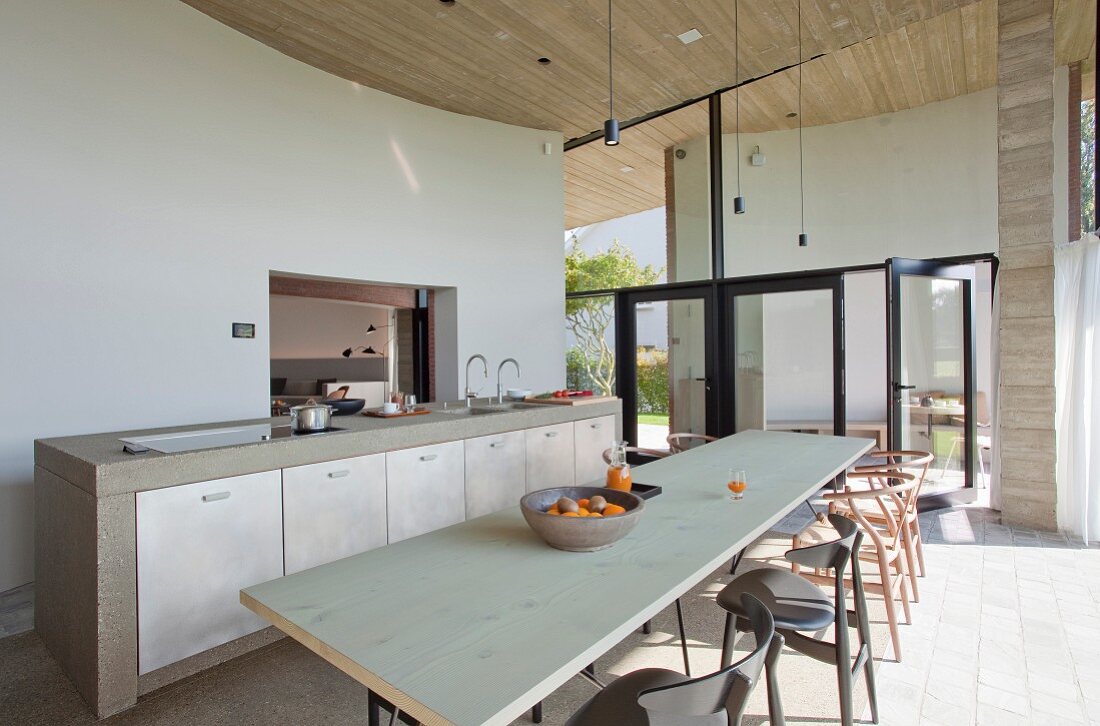 Long wooden table and classic chairs opposite masonry kitchen counter with pale cupboard doors in open-plan interior