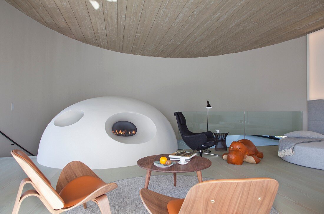 Extravagante Lounge in Kreisform davor massgefertigter Ofen mit Kaminfeuer als Halbkugel, im Vordergrund Klassikerstühle und Coffeetable aus Holz