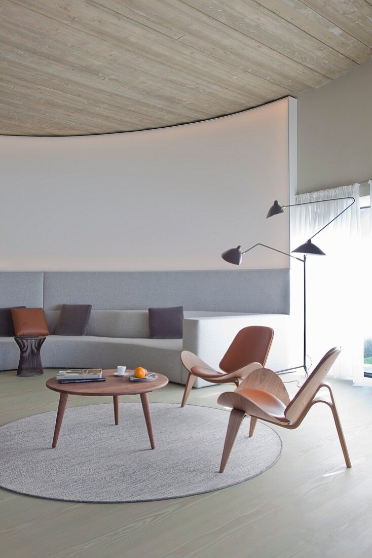 Klassikerstühle und Coffeetable aus Holz auf rundem Teppich vor massgefertigtem Sofa in Hellgrau, an gebogener Lounge Wand, seitlich Retro Stehleuchte