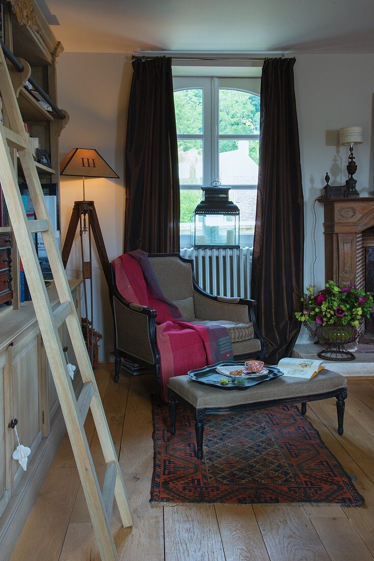 Wohnzimmer mit Holzleiter vor Bücherregal, im Hintergrund Stehleuchte neben Sessel am Fenster mit bodenlangem Vorhang