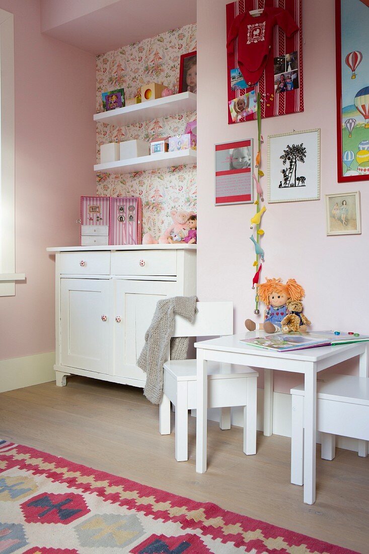 Child's bedroom with white furnishings, table and chairs against pink wall, chest of drawers and shelves in wallpapered niche