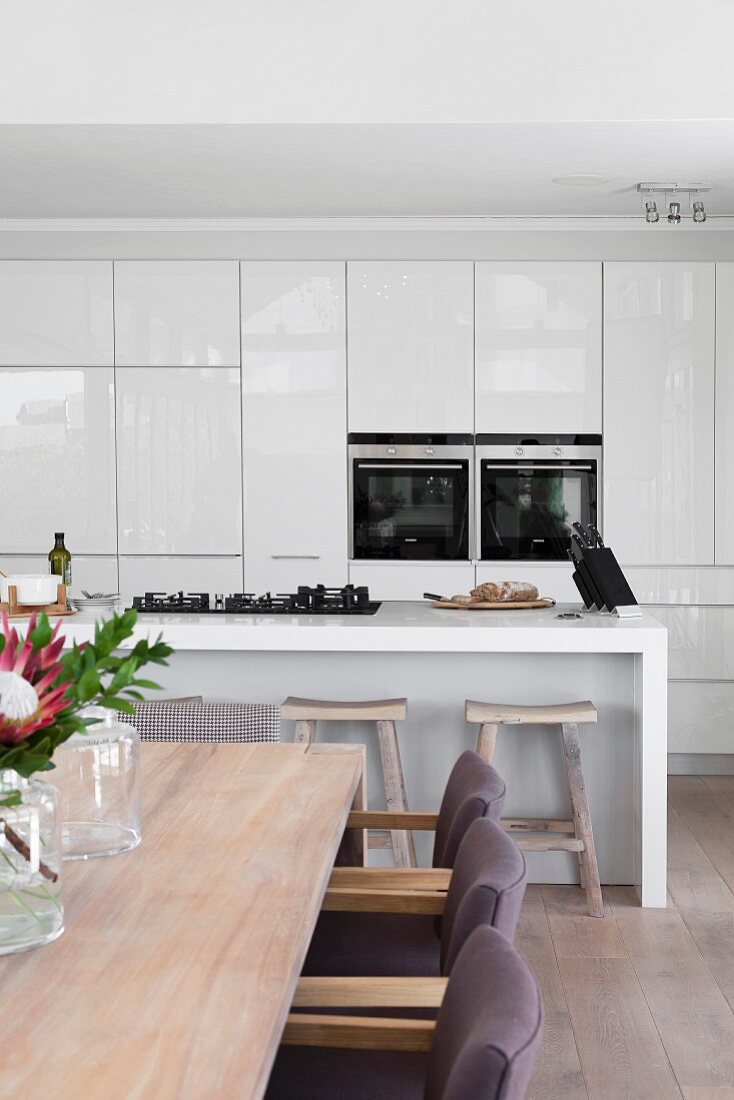 Ausschnitt eines Essplatzes vor offener Küche, minimalistische Theke mit Gasherd und Einbauschrank mit Geräten in hochglänzendem Weiß