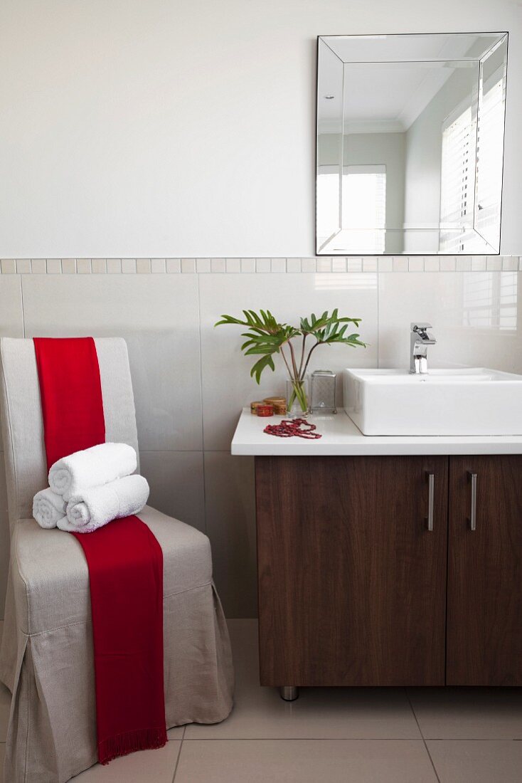 Gerollte Handtücher auf Hussensessel mit roter Schärpe, daneben Waschtisch mit Unterschrank und Aufsatzbecken