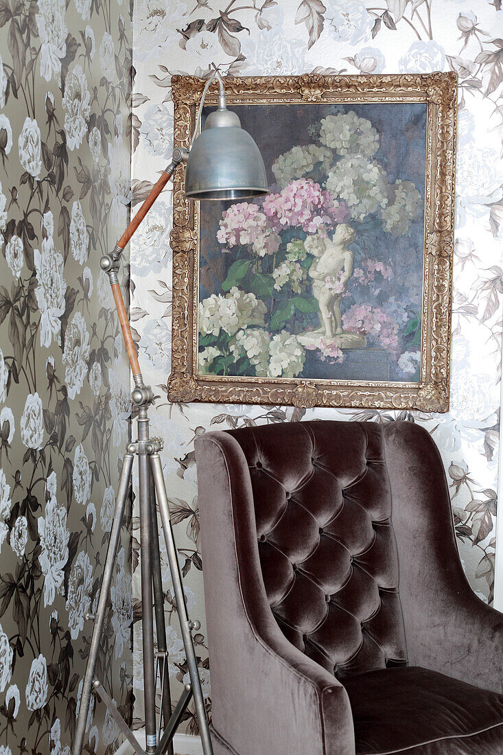 Brown velvet armchair and retro standard lamp below floral painting on floral wallpaper in corner