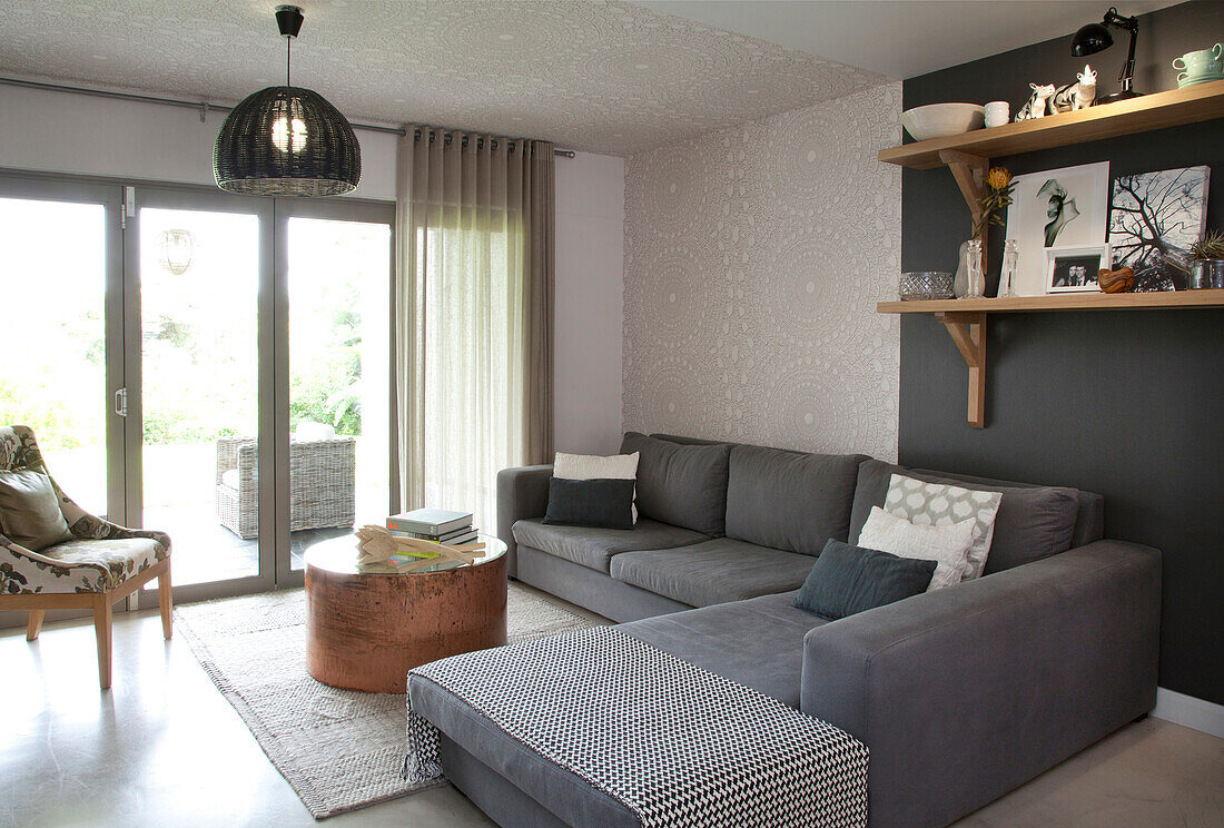 Moderne graue Sofalandschaft und runder Couchtisch in Wohnbereich mit Ornamenttapetenstreifen an Wand und Decke vor Terrassentüren