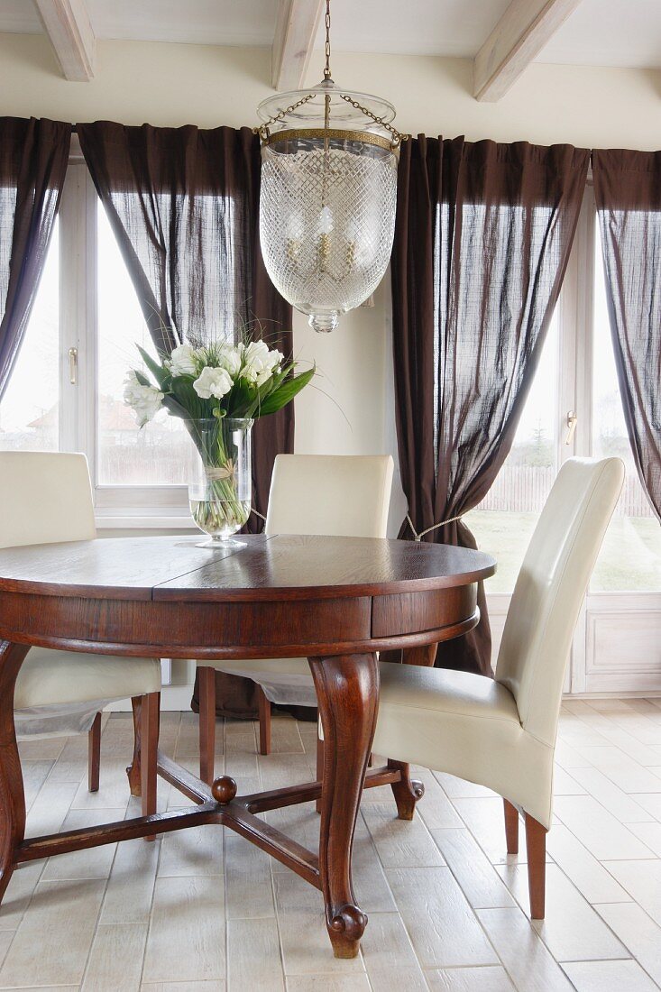 Traditionelle Hängeleuchte mit Glasschirm über rundem Holztisch im Biedermeier Stil und weiße, lederbezogene Stühle auf hellem Fliesenboden, im Hintergrund dunkle Gardinen am Fenster