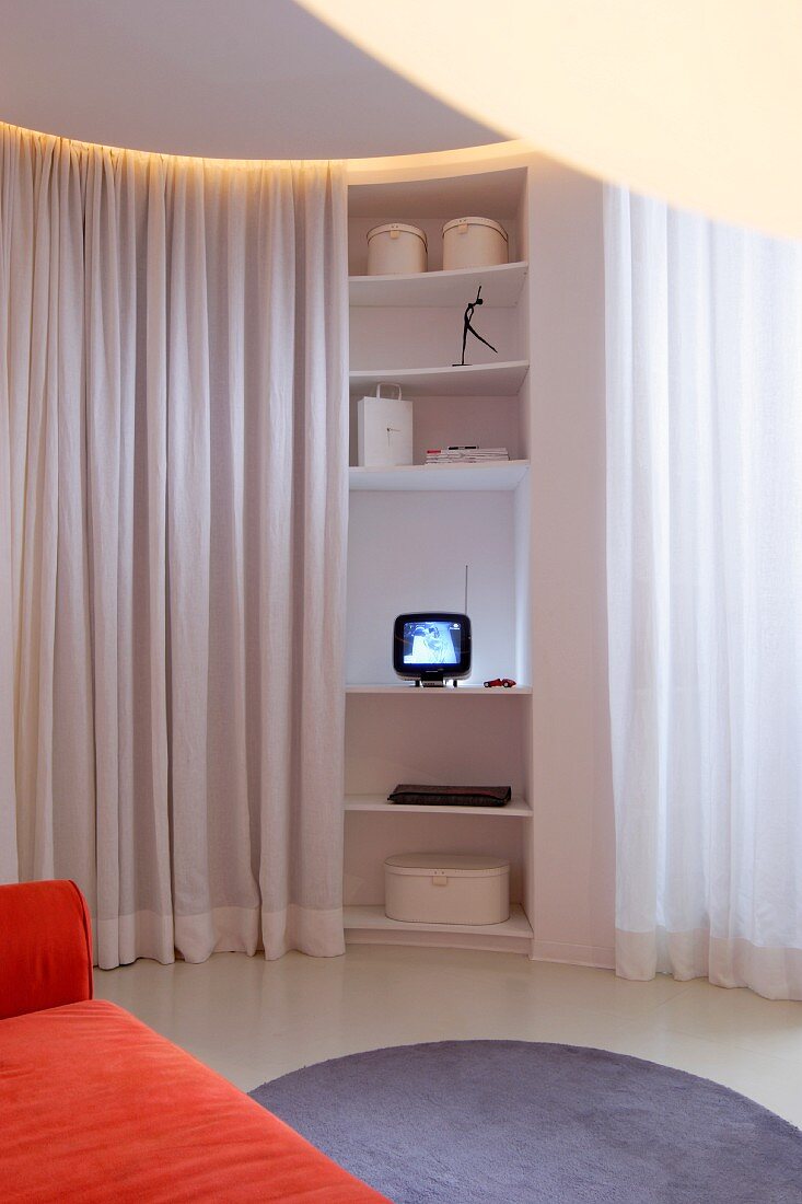 Geschwungenes Regal mit kleinem Fernseher zwischen weißem Vorhang als Raumabtrennung, runder hellgrauer Teppich vor orangefarbenem Sofa im Wohnbereich