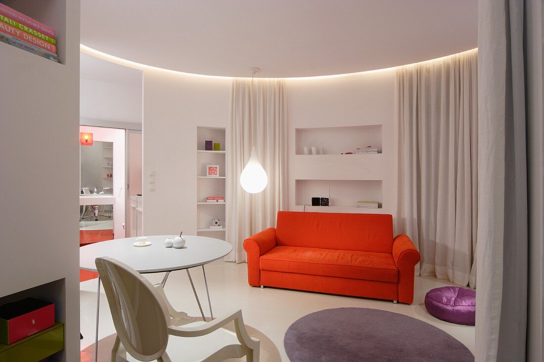 Orangefarbenes Sofa und weisser Tisch, Designer-Hängeleuchte in kreisförmigem modernem Appartement; weisser Vorhang zur Raumaufteilung