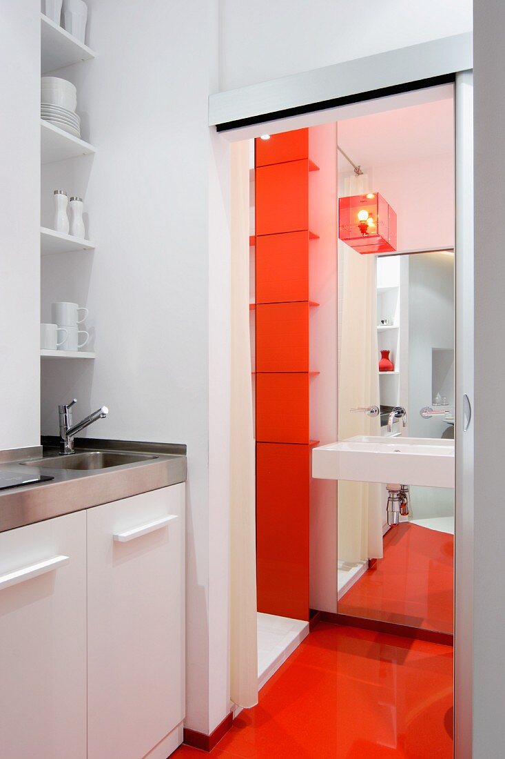 Miniküche mit Spüle und Regal in Nische, daneben offene Schiebetür mit Blick auf Waschbecken an raumhohem Spiegelpaneel und orangeroter durchgehender Boden