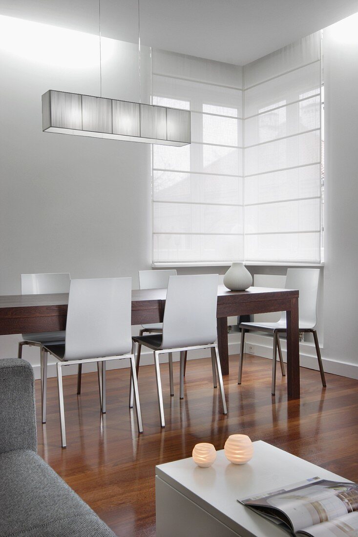 Stylische Wohnraumecke mit Essplatz, langer Holztisch und moderne Stühle auf edlem Holzboden, an Übereck-Fenster weiße, geschlossene Rollos