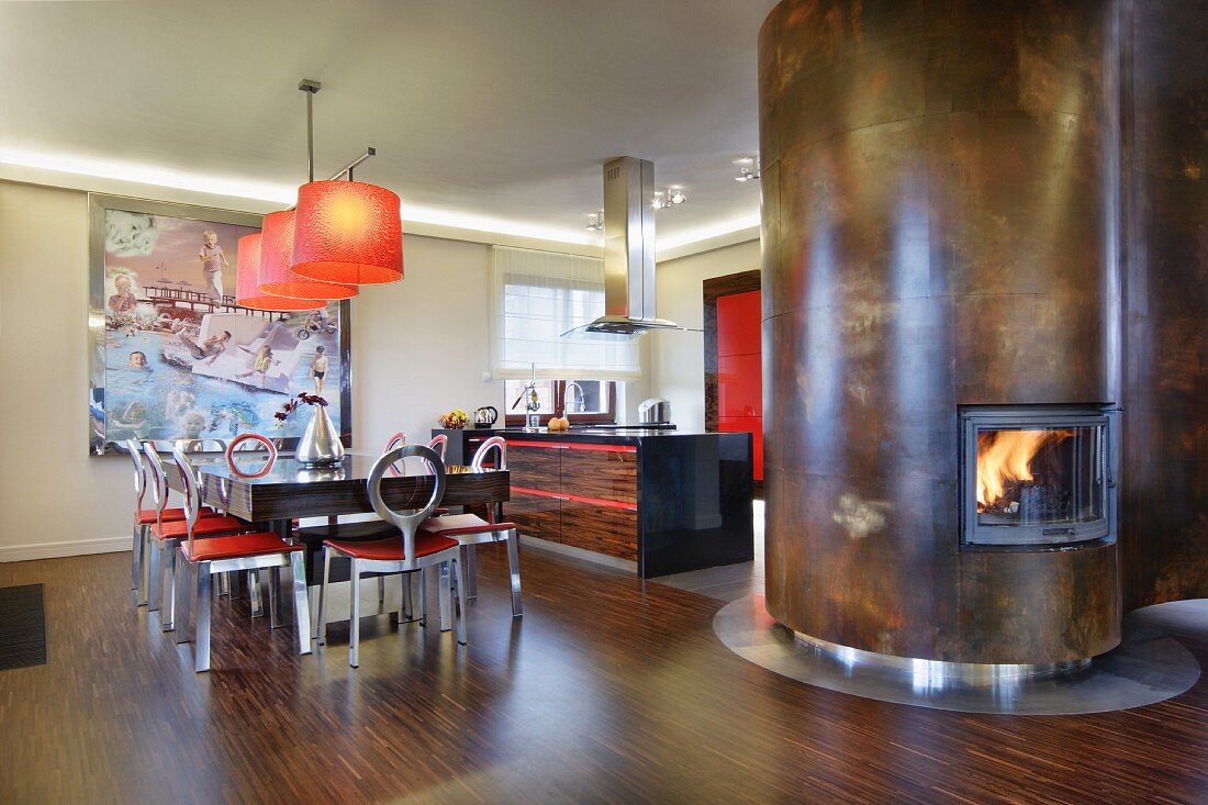Essplatz und offene Küche aus dunklem Holz, Edelstahl und roten Farbakzenten; geschwungener Raumteiler mit Metallverkleidung und integriertem Kaminofen