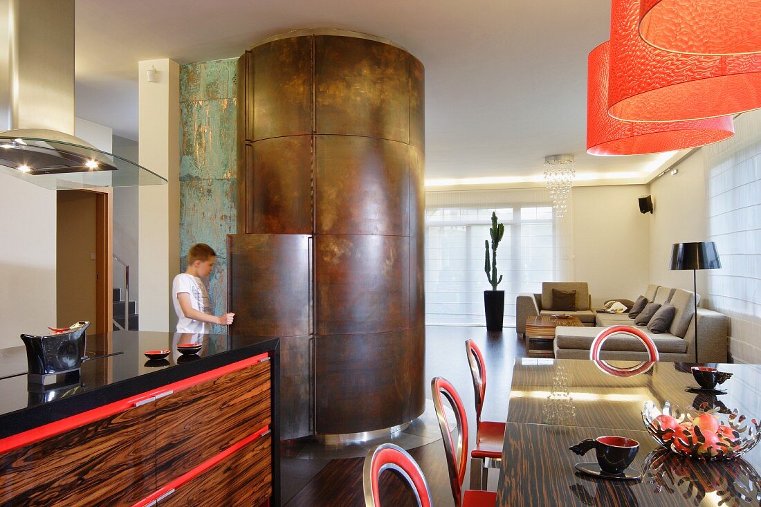 Dining area with varnished exotic wood surfaces and red accents; fridge in curved, sculptural partition with metal surface