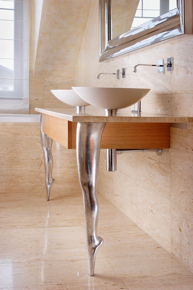 Twin sinks on wooden counter with stone slab surface and supports in shape of dancers legs en pointe in bathroom with stone wall and floor tiles
