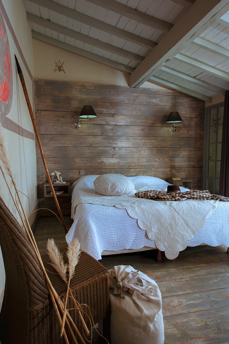 White bedspread on double bed below sconce lamps on wood-clad wall in rustic bedroom
