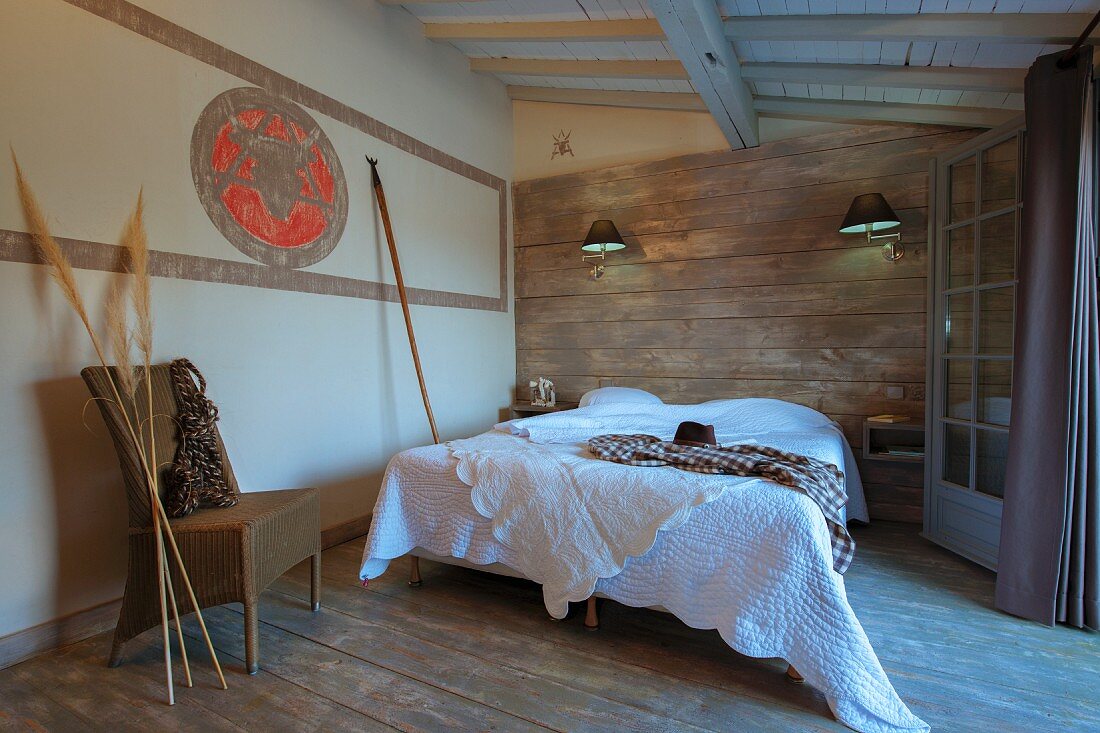 Double bed with white bedspread against wood-clad wall, wicker chair and pampas grasses to one side in rustic bedroom