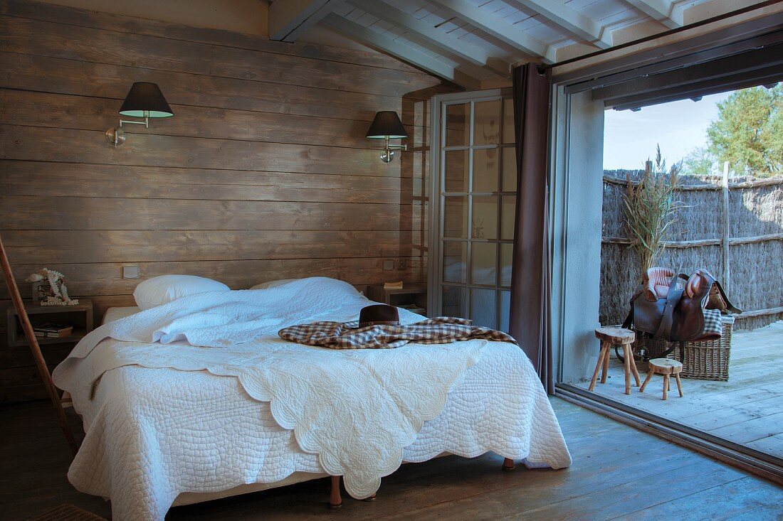 Doppelbett mit weisser Tagesdecke vor holzverschalter Wand in rustikalem Schlafzimmer, seitlich offene Terrassentür