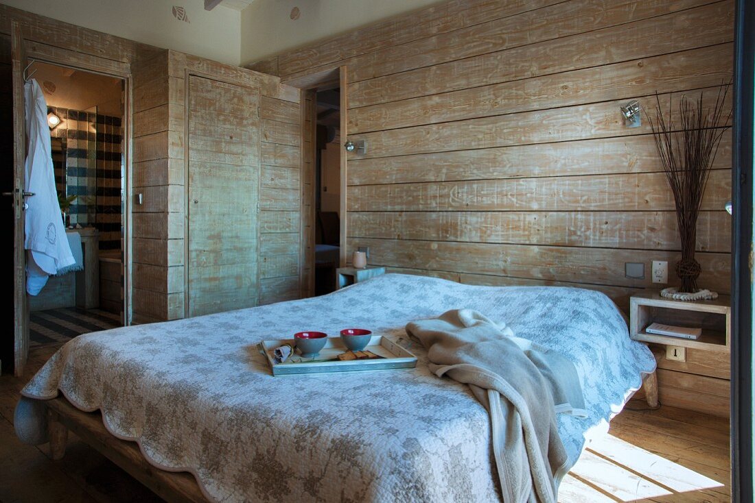 Breakfast tray on double bed with bedspread in rustic bedroom with horizontal wooden cladding
