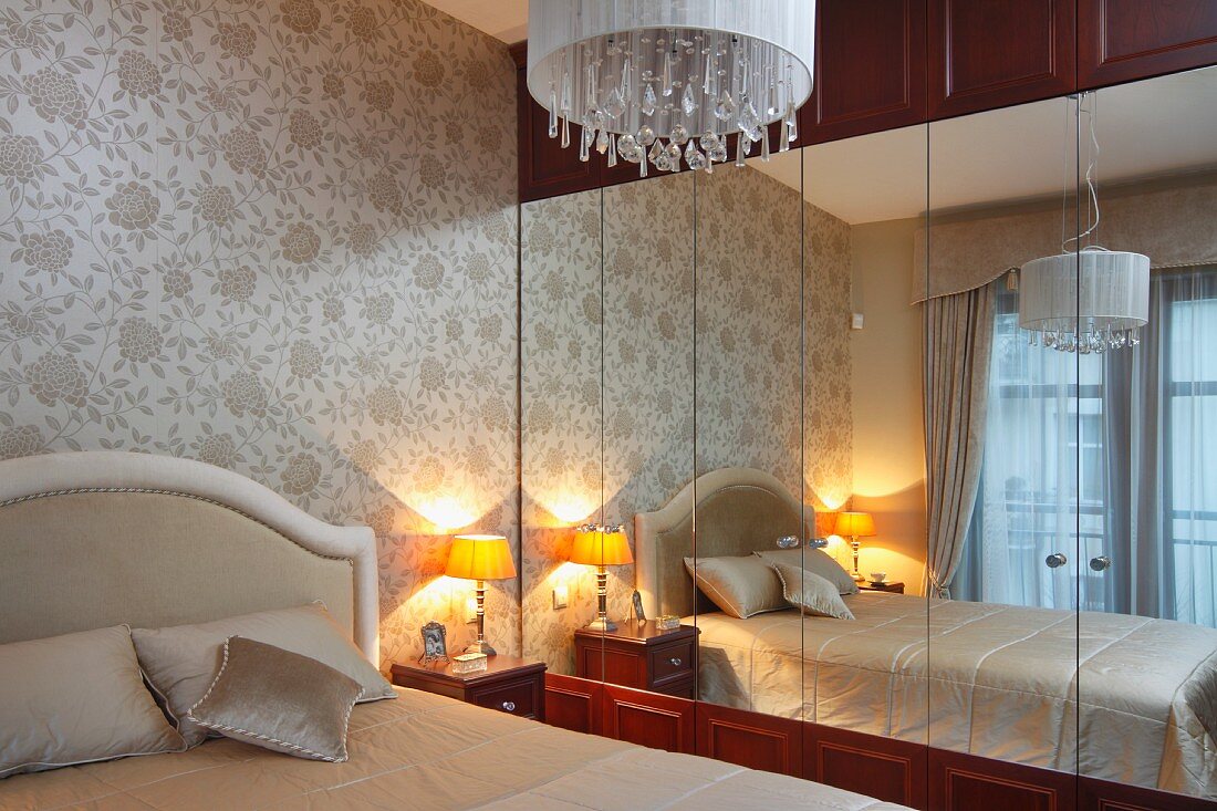 Ausschnitt eines Bettes mit Polster Kopfteil und Tischleuchte auf Nachtschränkchen neben Einbau Spiegelschrank