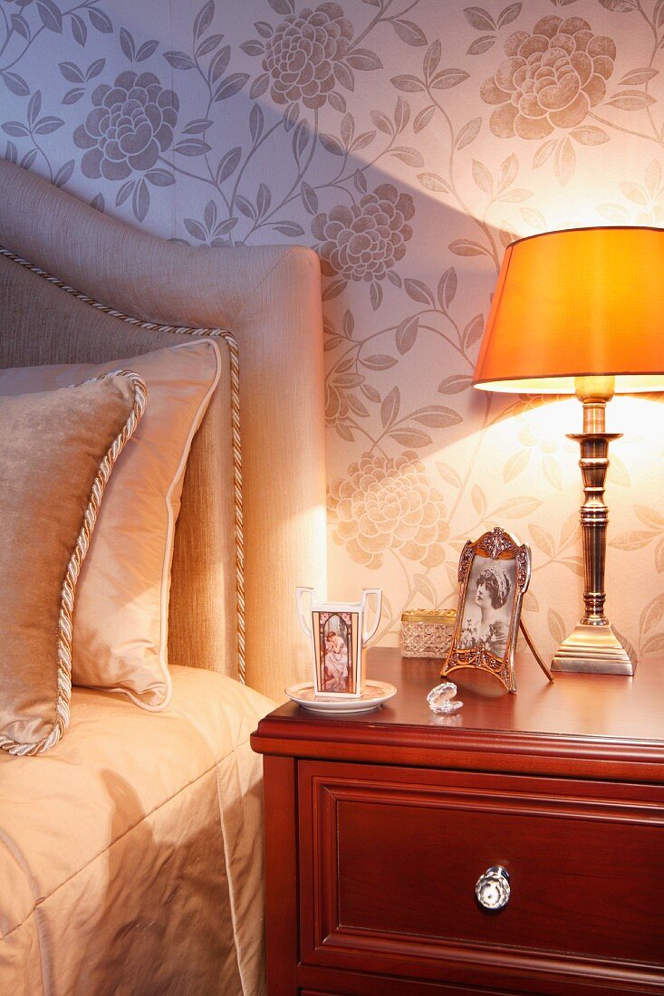 Bedside lamp with yellow lampshade on mahogany bedside cabinet; bed with headboard against wall with pale, silk floral wallpaper
