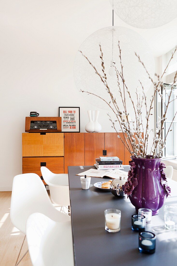 Weidenzweigen in violetter Vase und Windlichter auf Tisch, davor weiße Klassikerstühle