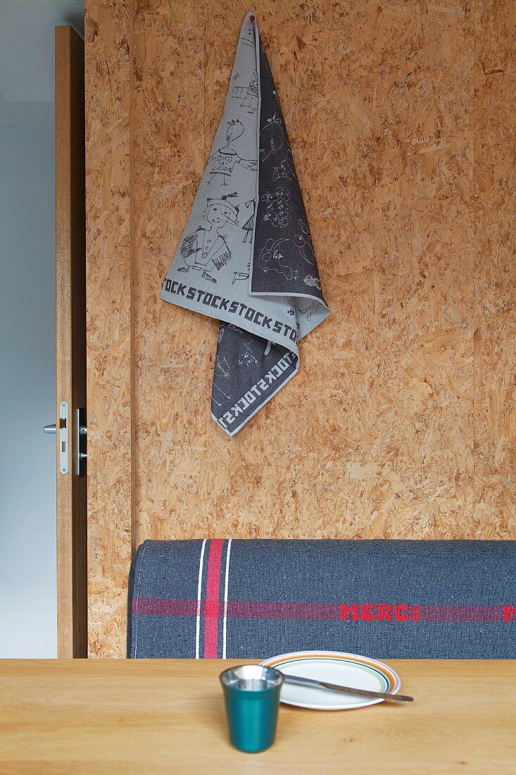 Printed tea towel pinned to wall made from plain OSB board above grey backrest of bench and place setting on dining table