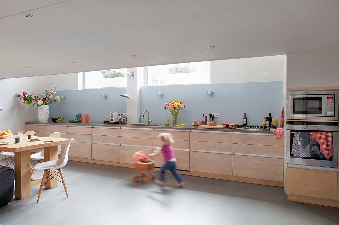 Langgestreckte Küchenzeile mit hellen Holzfronten und hellblauem Spritzschutz in Wohnküche mit spielendem Mädchen