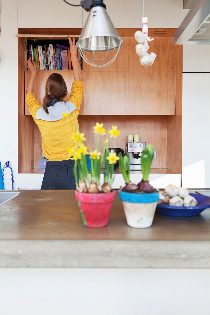Blumentöpfe mit Narzissen und Hyazinthen auf Küchentheke, im Hintergrund Frau vor Bücherschrank aus Holz