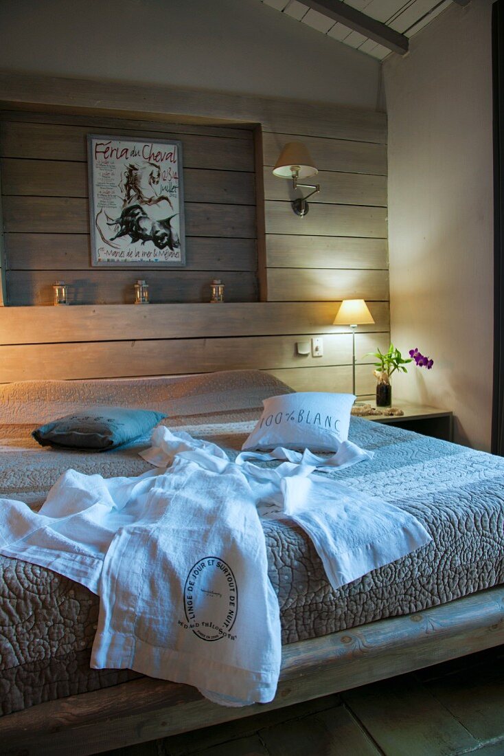 Weisser Morgenmantel auf Bett, vor holzverschalter Wand mit Nische, seitlich Tischleuchte